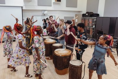 Women drummers from Rwanda teach their drumming techniques to a group of 澳门六合彩平台网站投注 students. The musicians and the students stand in the middle of a room, circling six big barrel drums, many of them holding drumsticks high in the air, ready to make contact with the drums. The drummers are are Black women with brightly colored long braids.  They wear multi-colored patterned dresses. The students are dressed in sweatshirts and jeans and sweatpants.